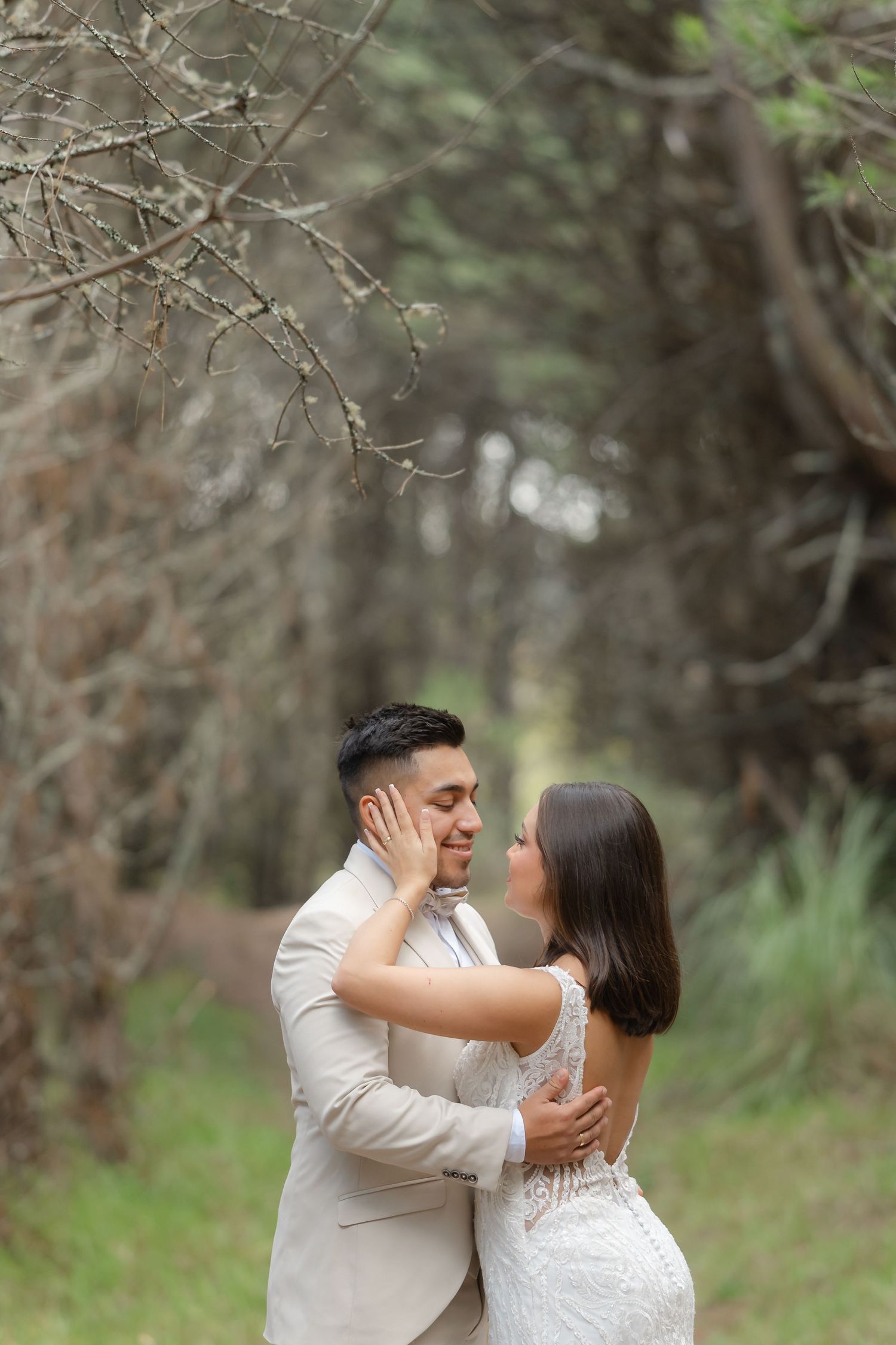 A couple in white attire shares an intimate moment while embracing in a wooded outdoor setting.
