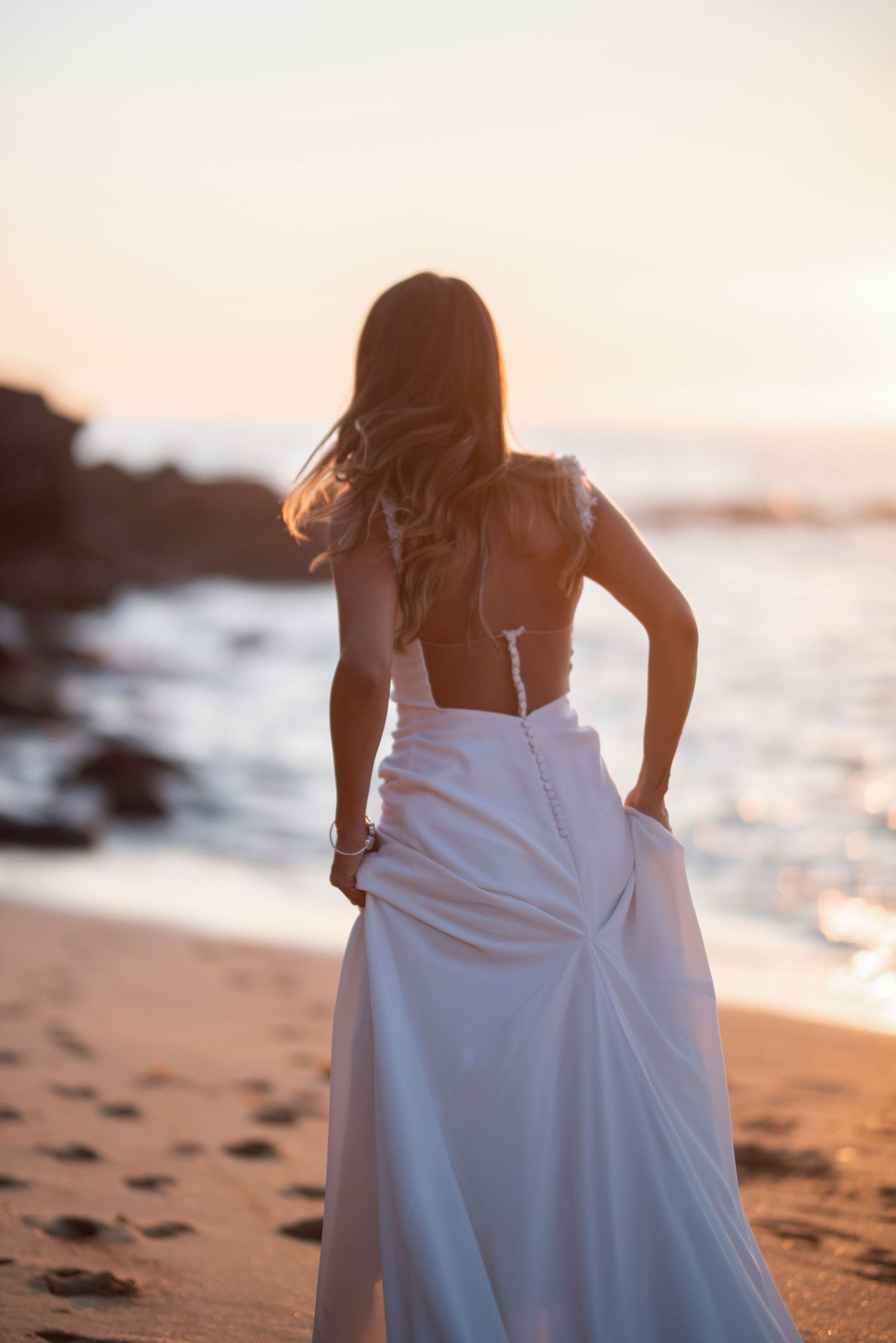 A woman in a flowing white dress walks along a beach at sunset with waves crashing on rocky shore.