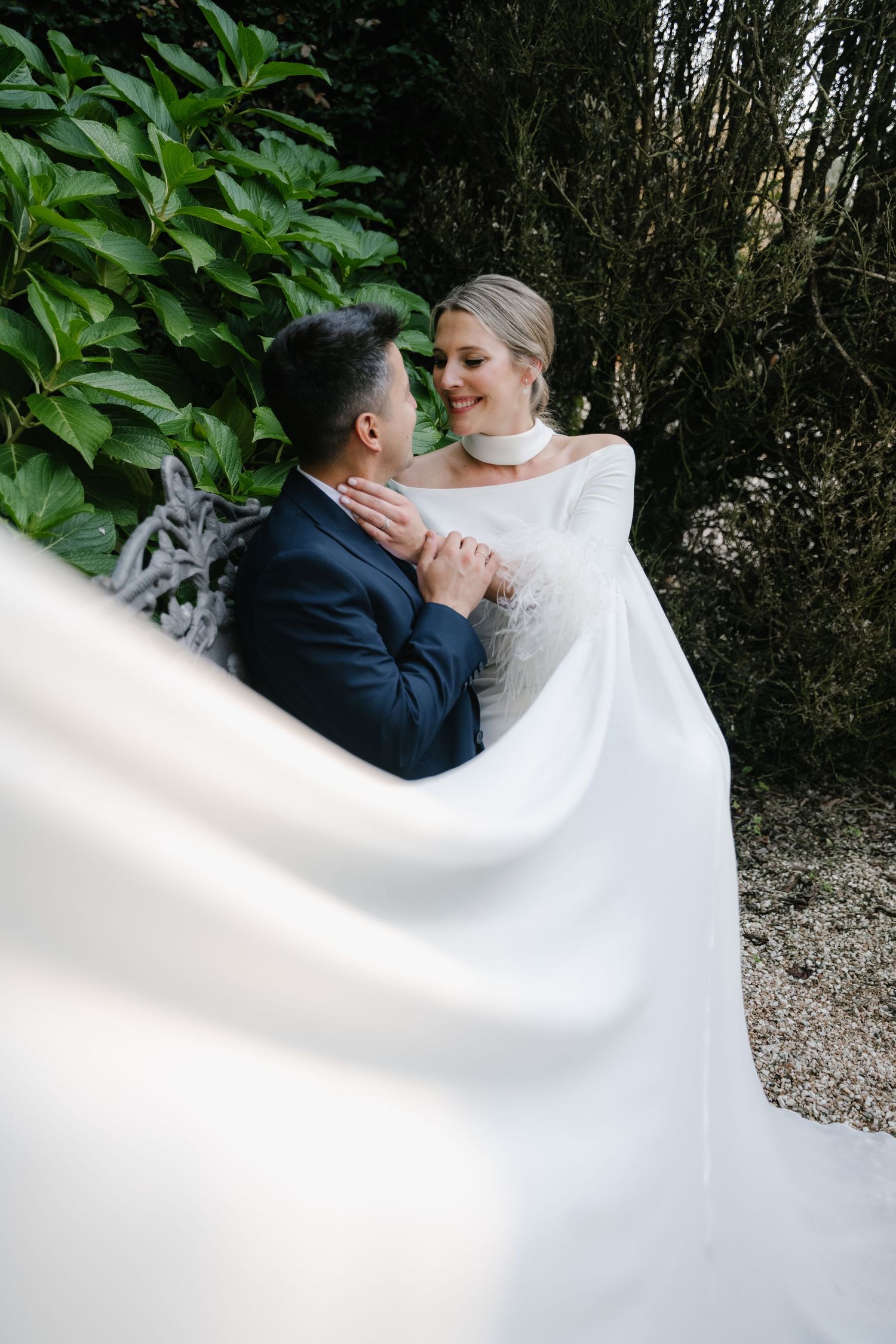 A romantic wedding couple embraces while standing in a garden with lush greenery in the background.