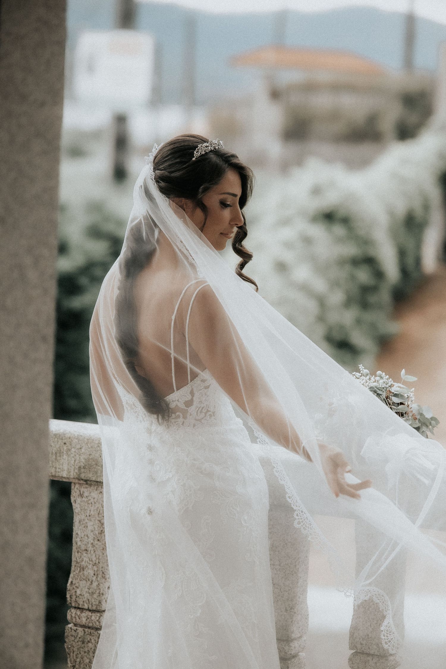 A bride in a flowing white wedding dress and veil poses elegantly against stone pillars in a romantic outdoor setting.