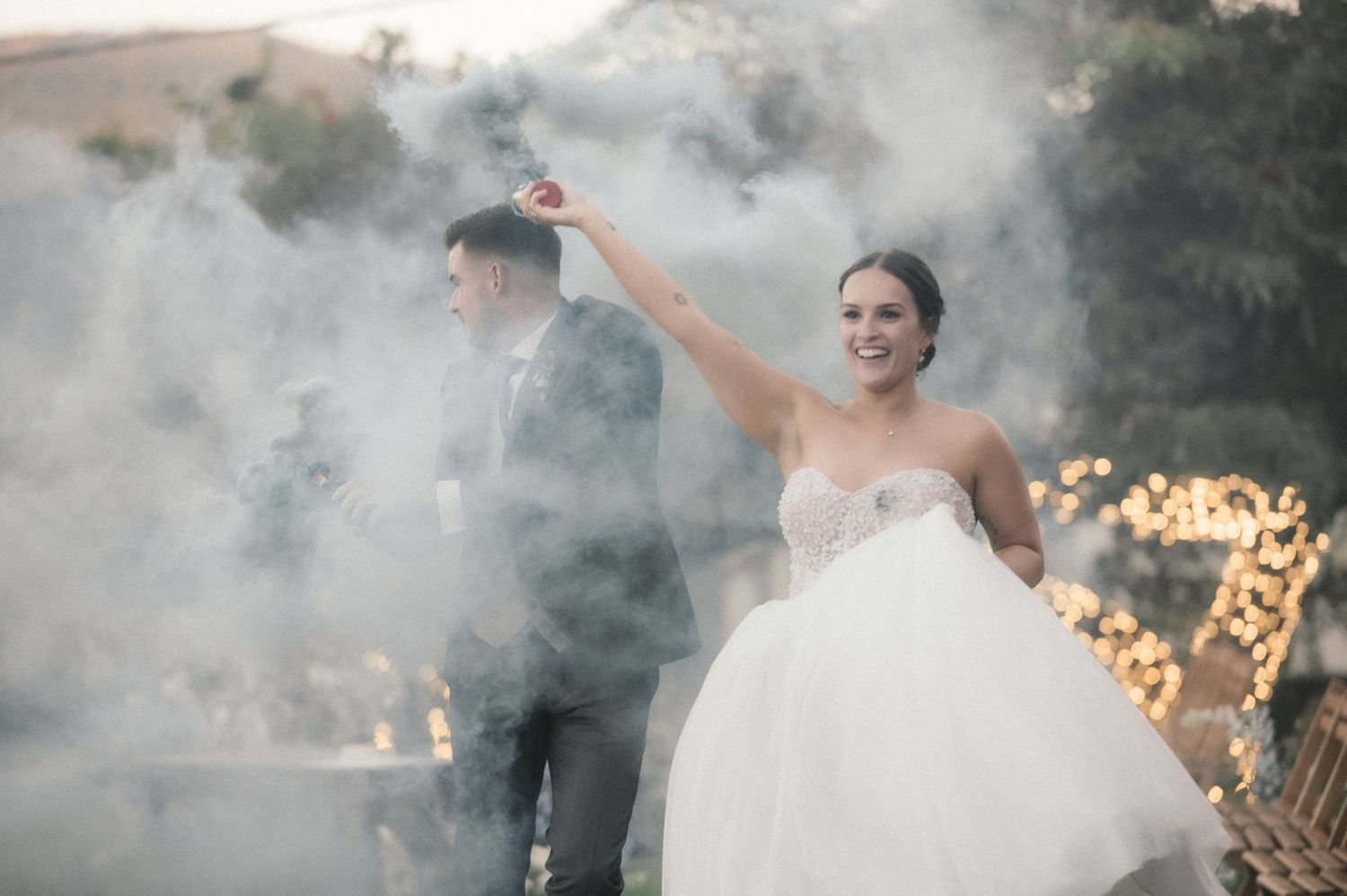 Wedding party celebrates with smoke effects and string lights creating a dreamy outdoor atmosphere.