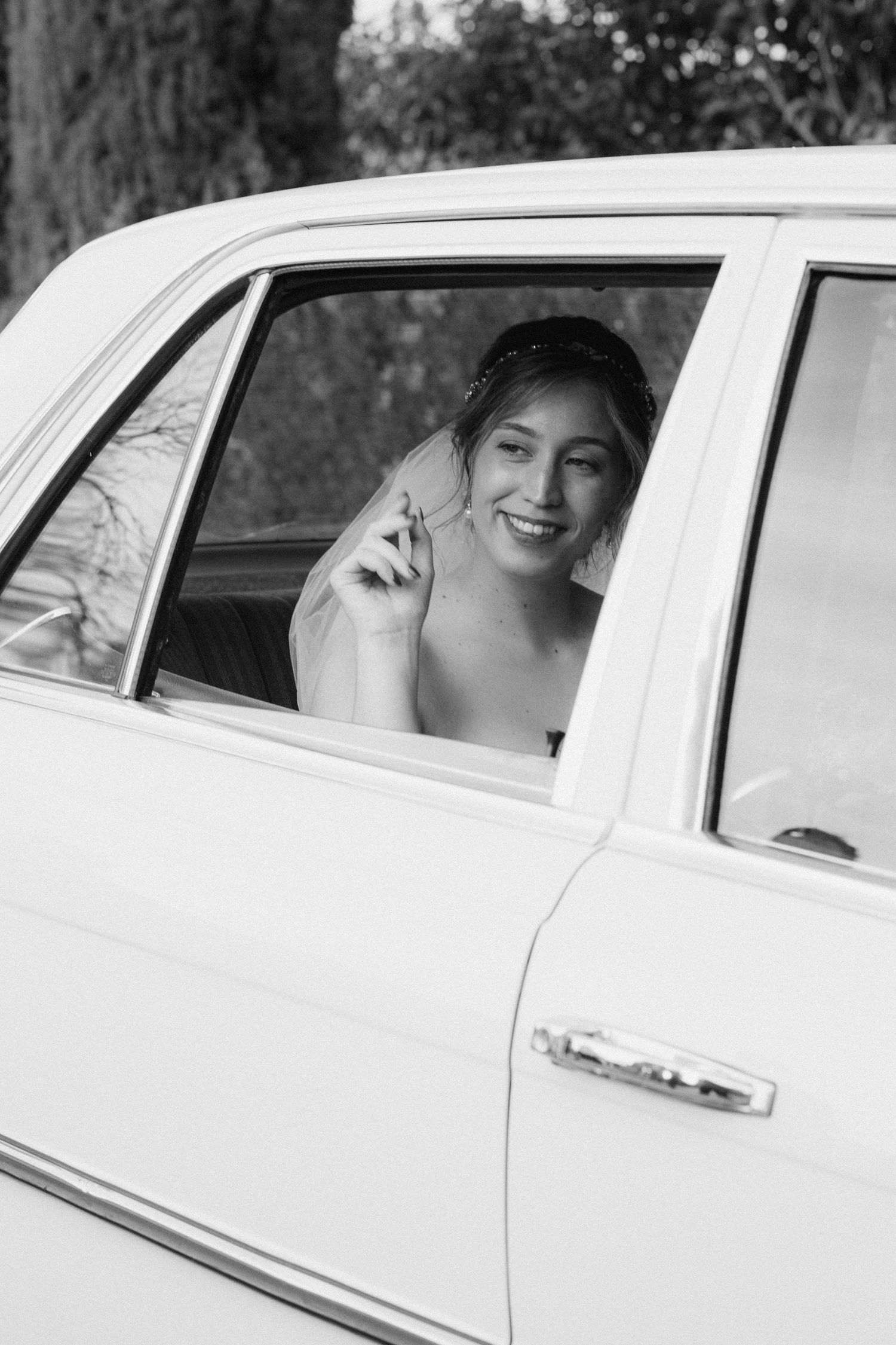 A black and white photograph shows someone smiling through a car window of a classic white automobile.