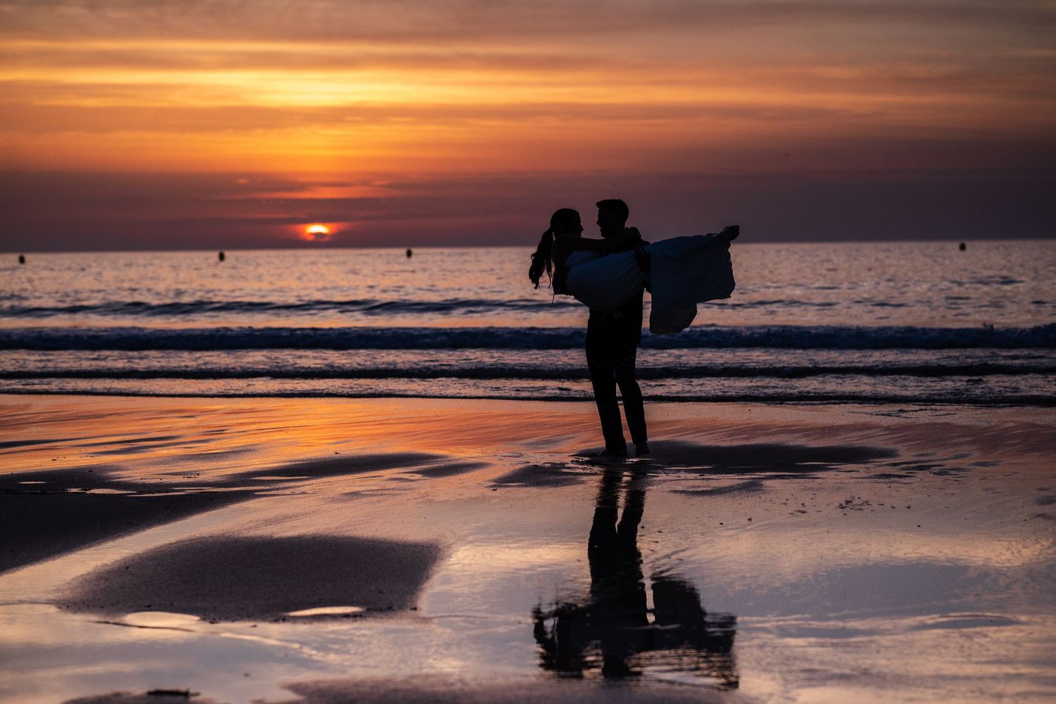 Silhouettes of beachgoers enjoying a vibrant orange sunset over the ocean waves.