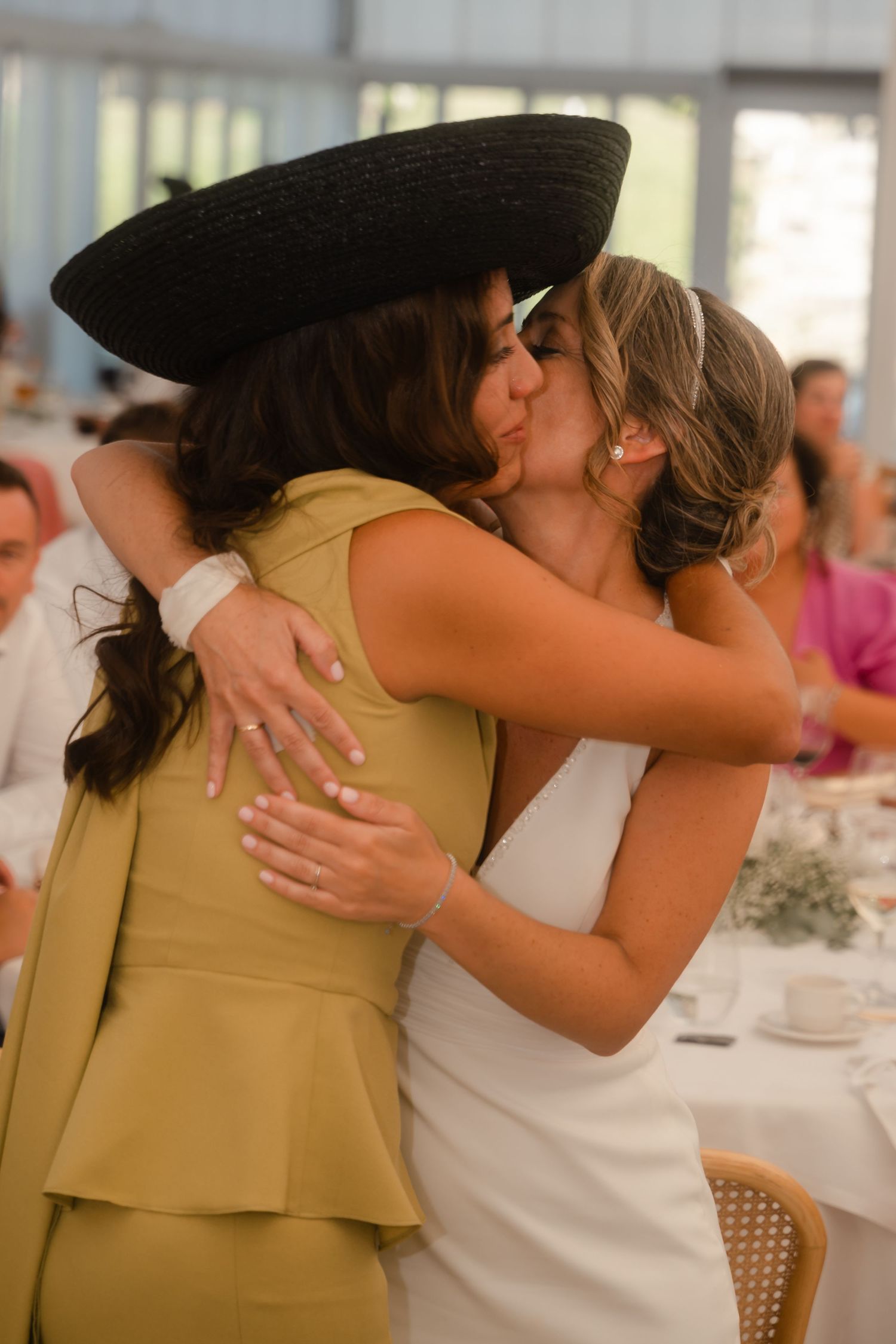 Two wedding guests share an emotional embrace at a reception, one wearing a yellow dress with large hat and one in white.