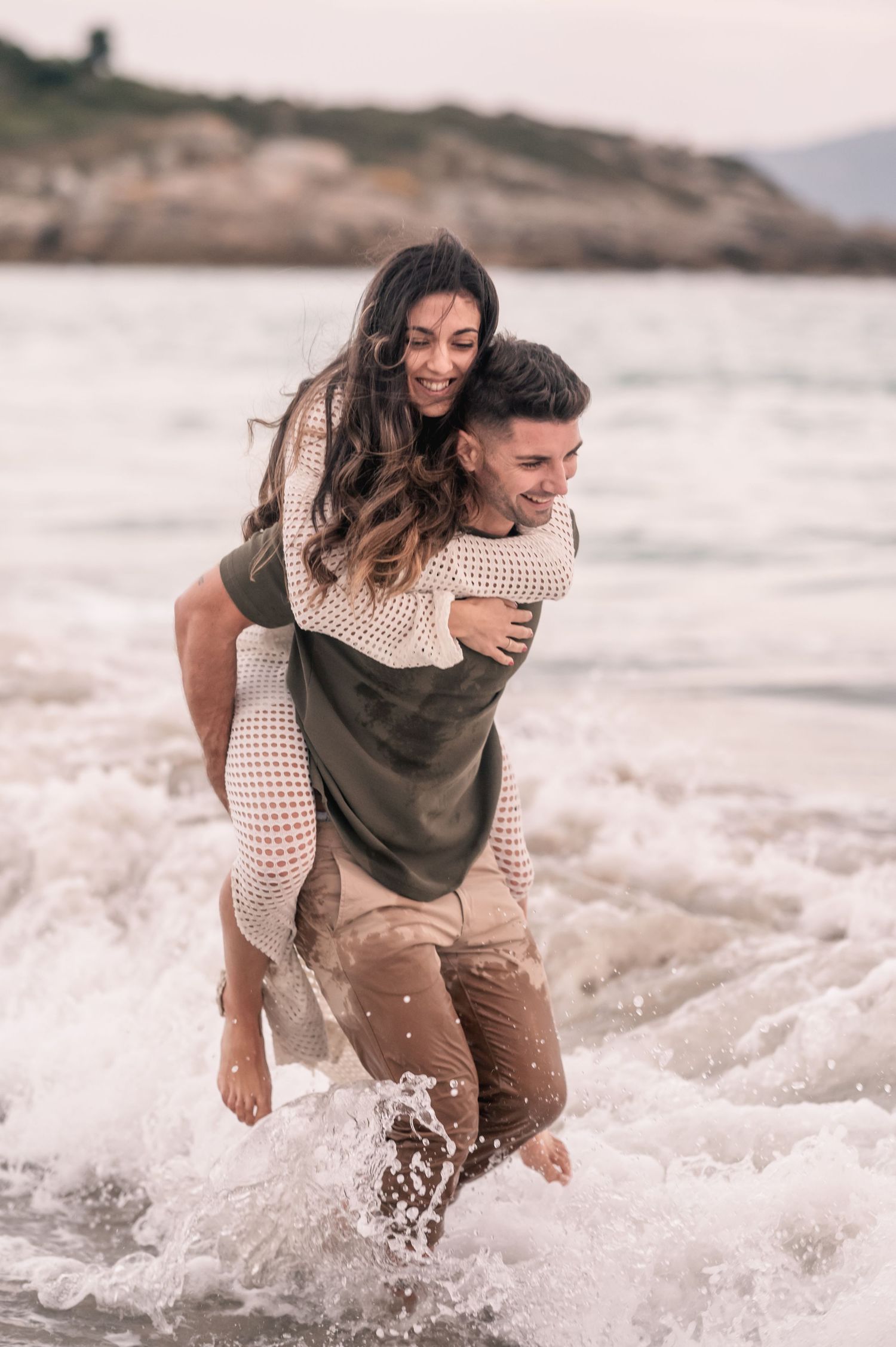 A couple plays in the ocean waves at sunset, with one person giving a piggyback ride while splashing through the water.