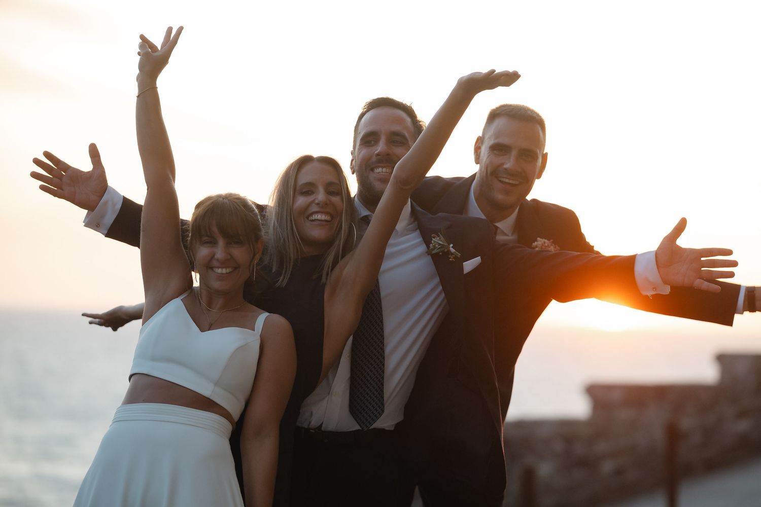 Group of friends celebrating together on a beach at sunset with arms raised in joyful expressions.