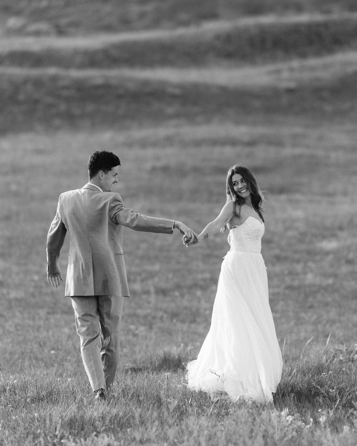 Black and white photo of a bride and groom holding hands in a grassy field on their wedding day.