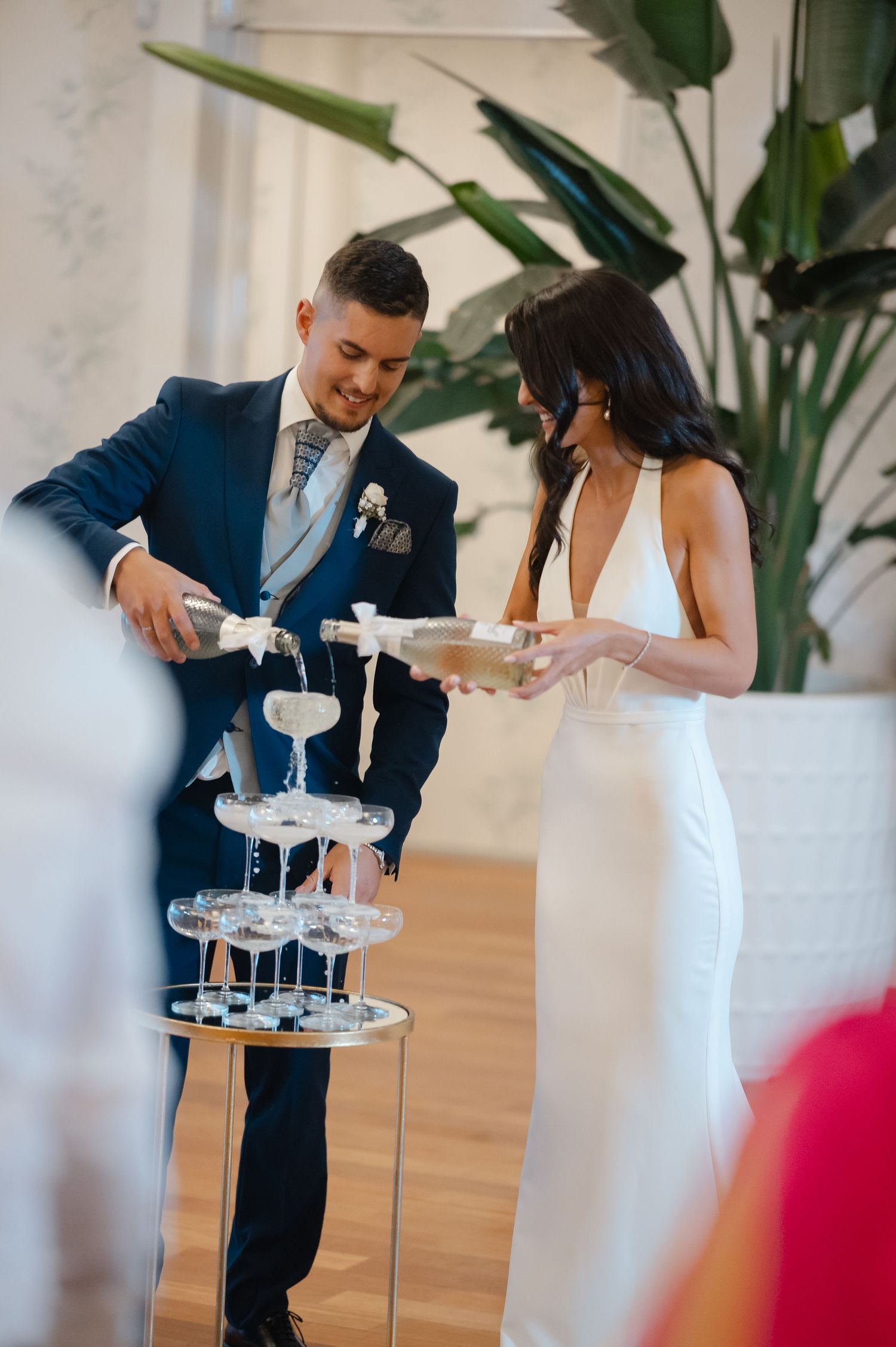Couple in formal attire pouring champagne into a tower of glasses at an elegant celebration.