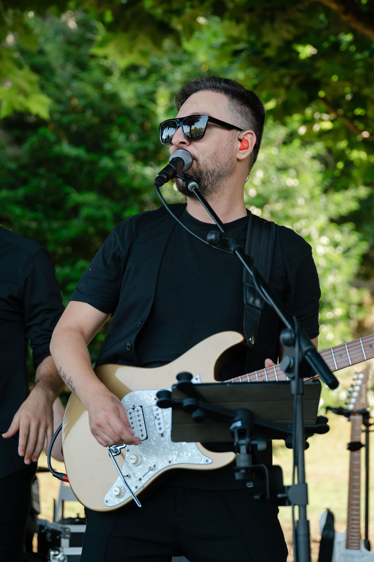 Musician in black clothing performs with an electric guitar and microphone in an outdoor setting with green foliage.