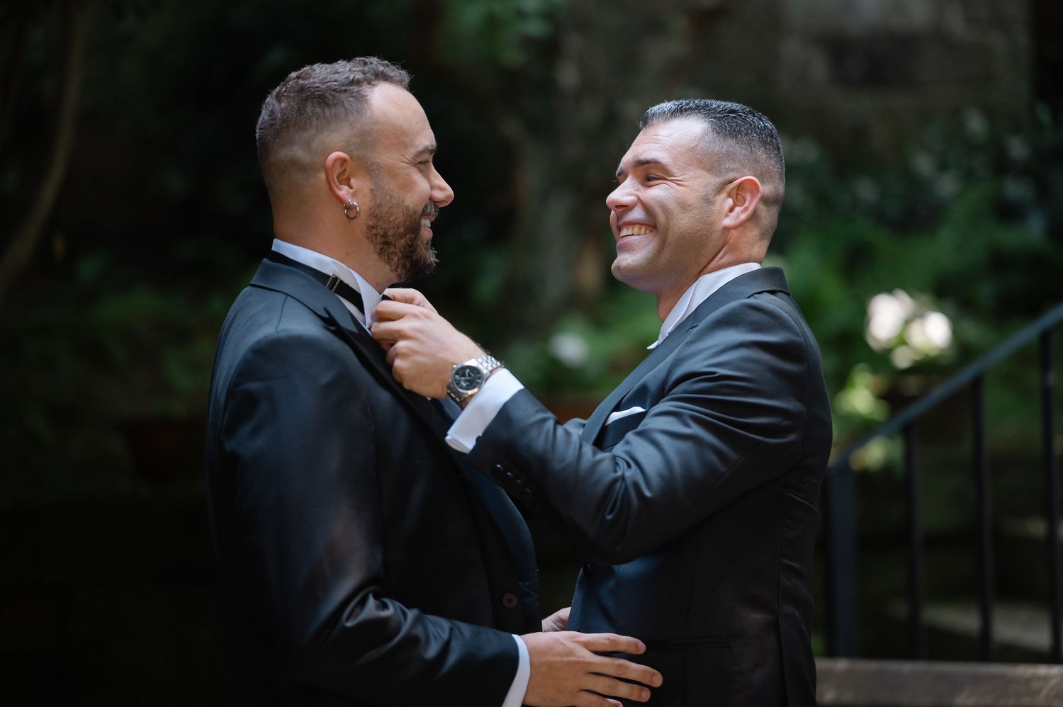Two people in formal suits embrace warmly while sharing a genuine laugh together outdoors.
