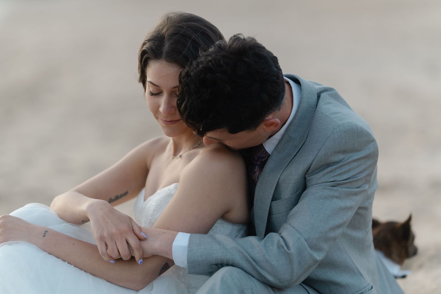 A groom in a gray suit embraces his bride in a white dress on a beach during a romantic wedding photo shoot.