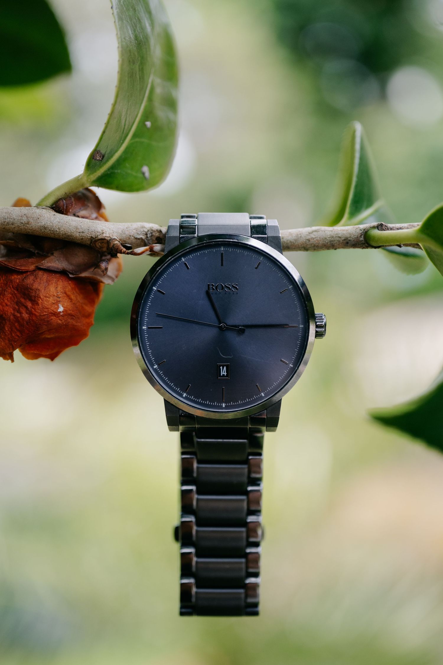 A sleek black minimalist wristwatch with metal bracelet photographed against a blurred natural green background.