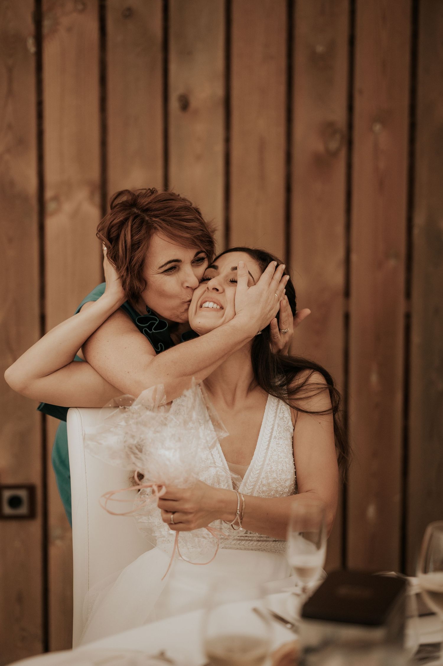 Two people share a joyful embrace in front of a rustic wooden wall during a wedding celebration.