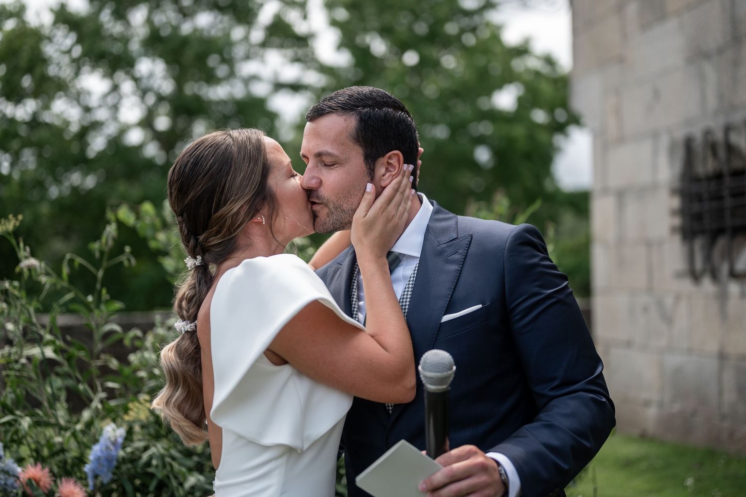 Couple shares romantic kiss during outdoor wedding ceremony with stone building backdrop.