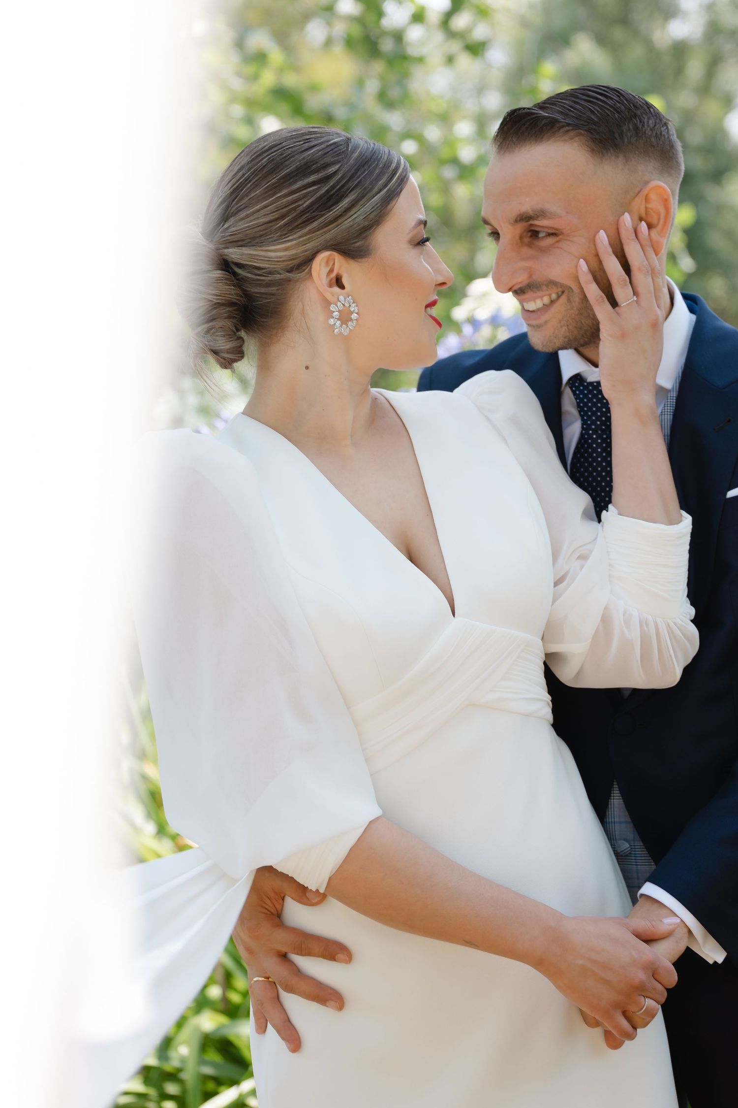 A romantic wedding photo of a couple in white dress and navy suit sharing an intimate moment in a garden setting.
