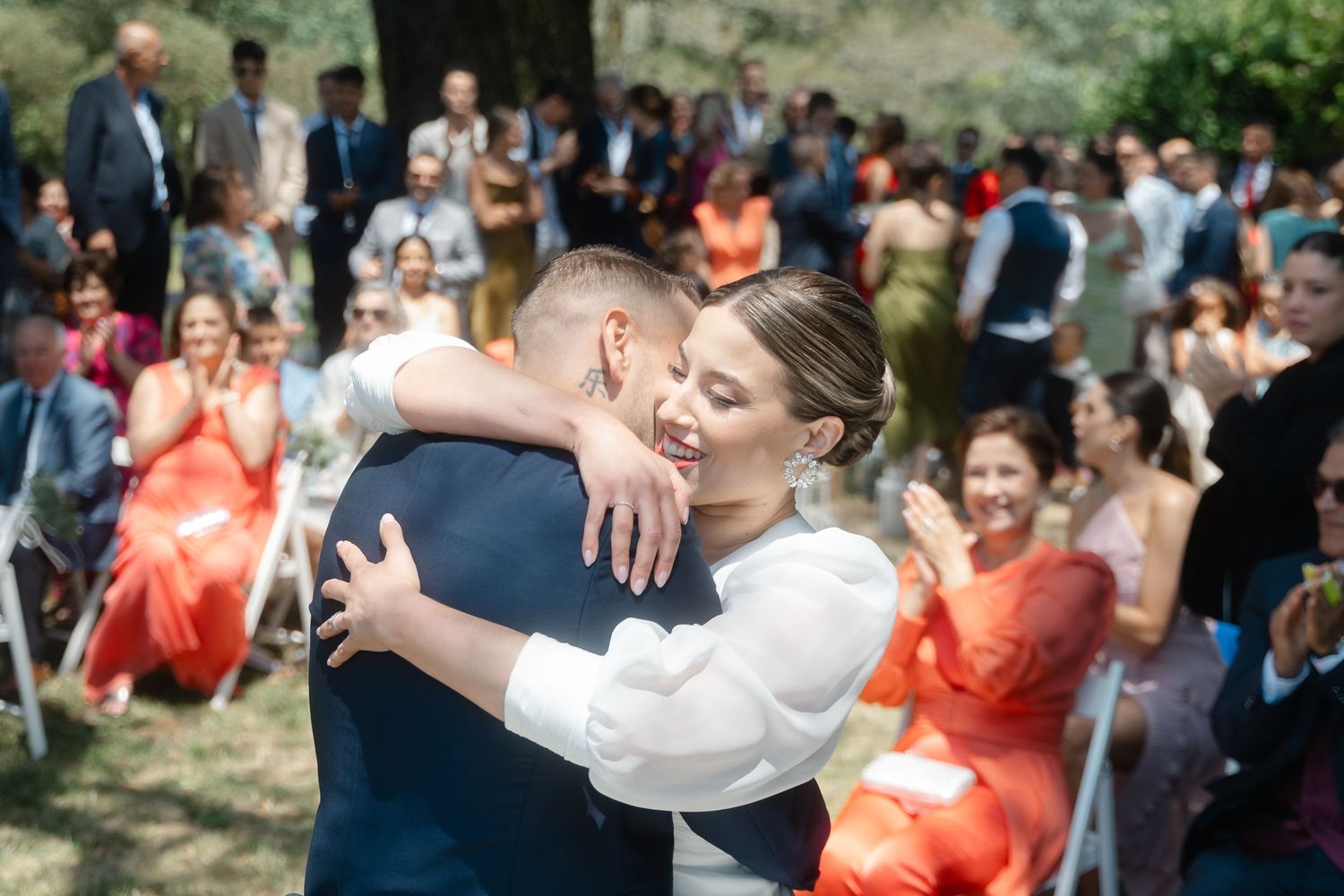 Wedding party members in coral dresses celebrate with joyful embraces during outdoor reception festivities.