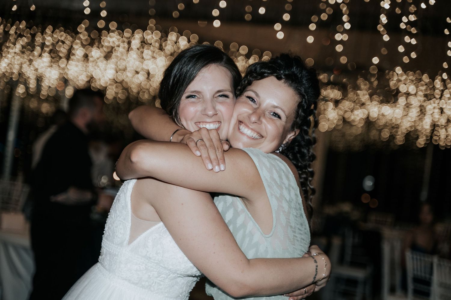 Two people share a joyful embrace at a wedding reception under twinkling string lights.