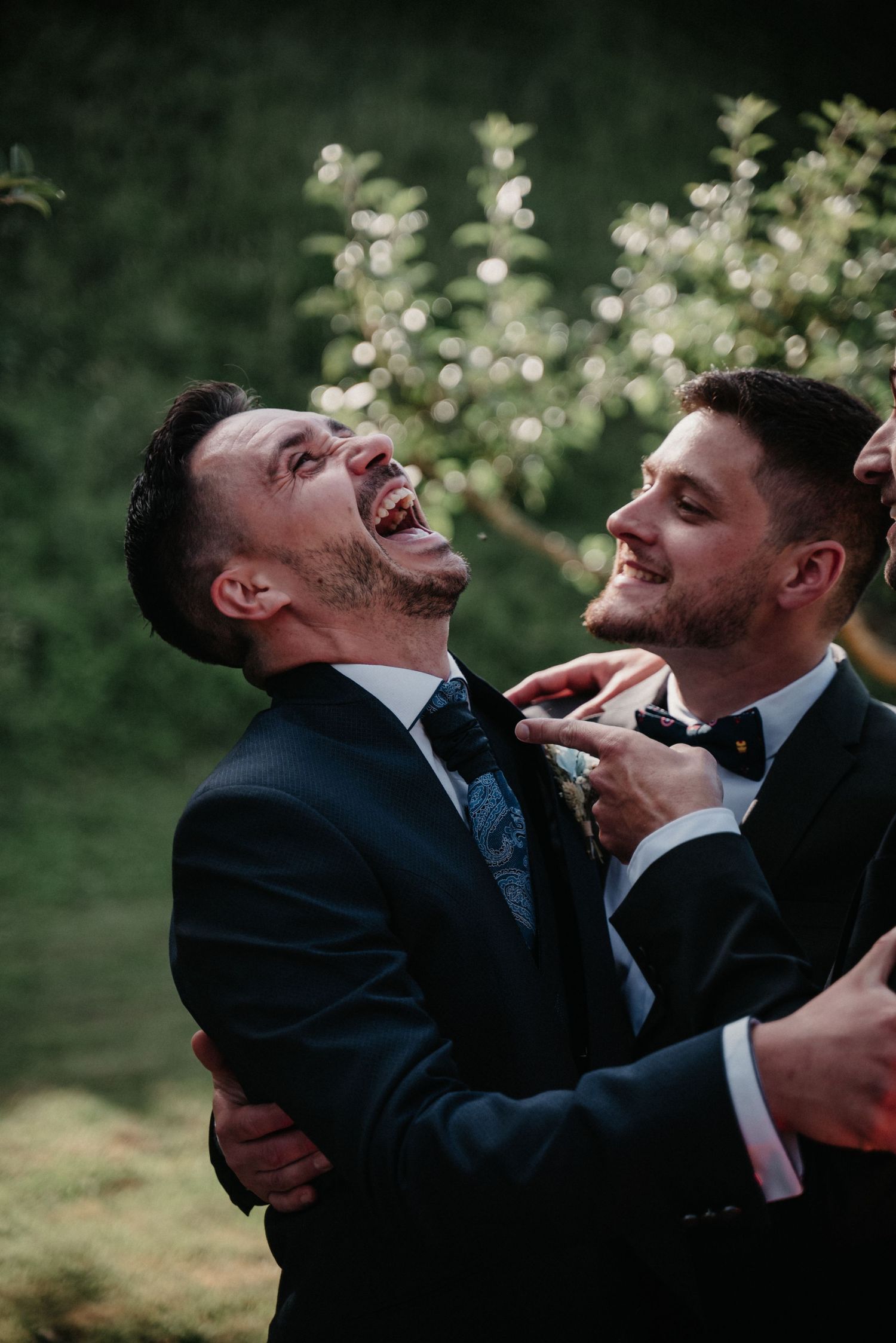 Two groomsmen in dark suits share a joyful candid moment while laughing together outdoors.