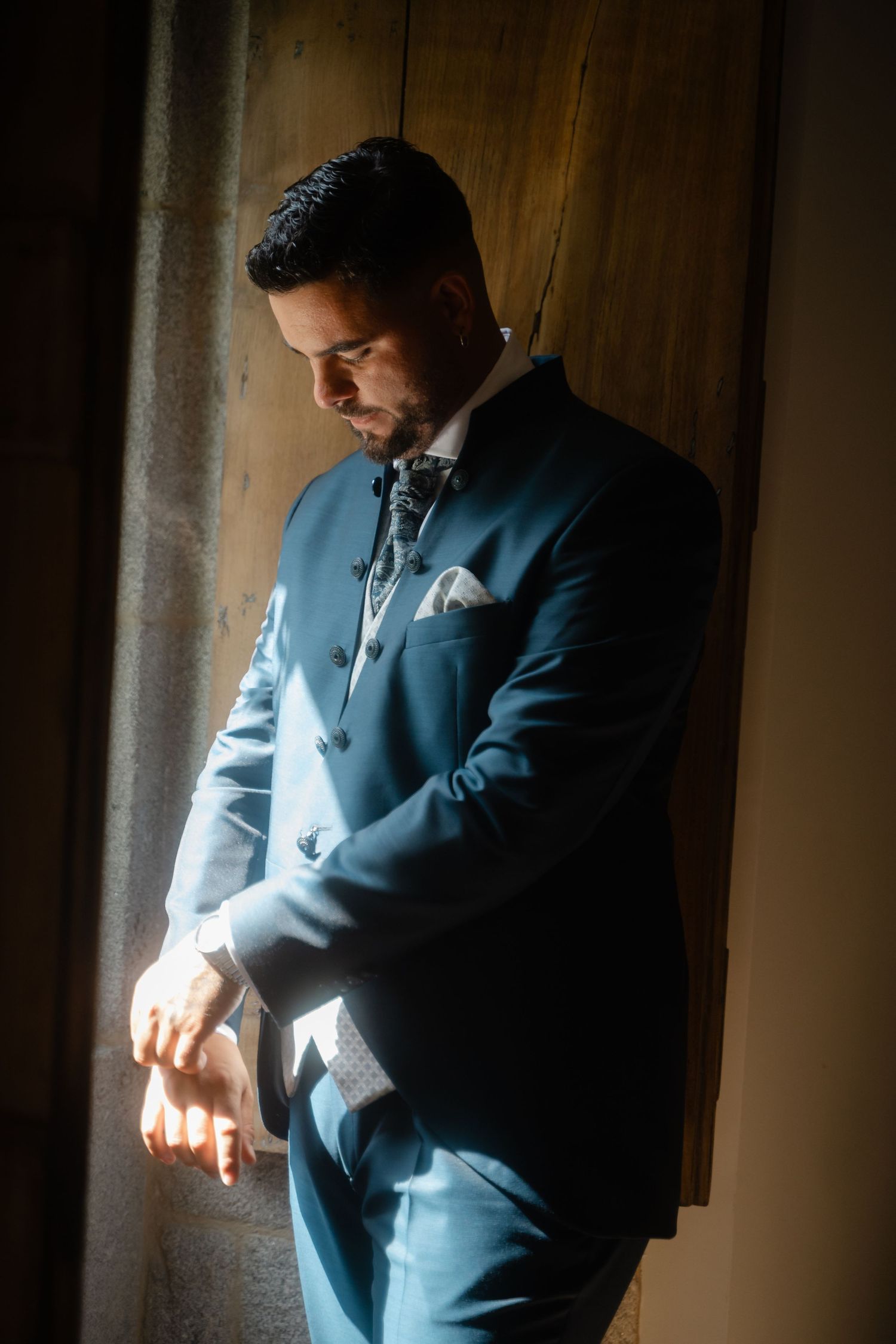 A person in a stylish blue suit adjusts their cuffs while standing in dramatic window lighting.