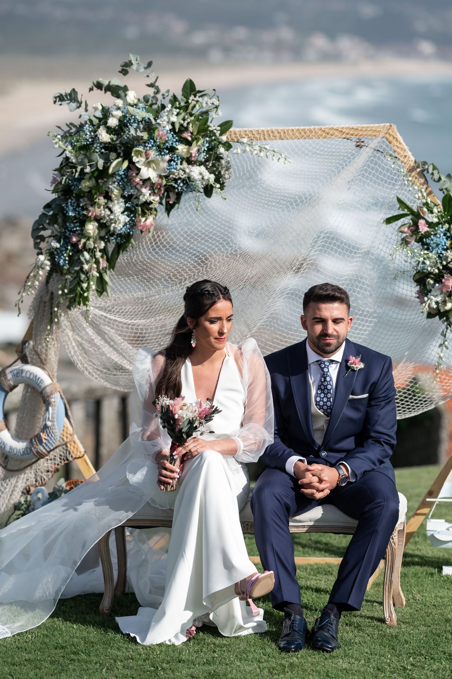 A bride's veil flows dramatically in the wind during outdoor wedding photos with her groom on a grassy hillside.