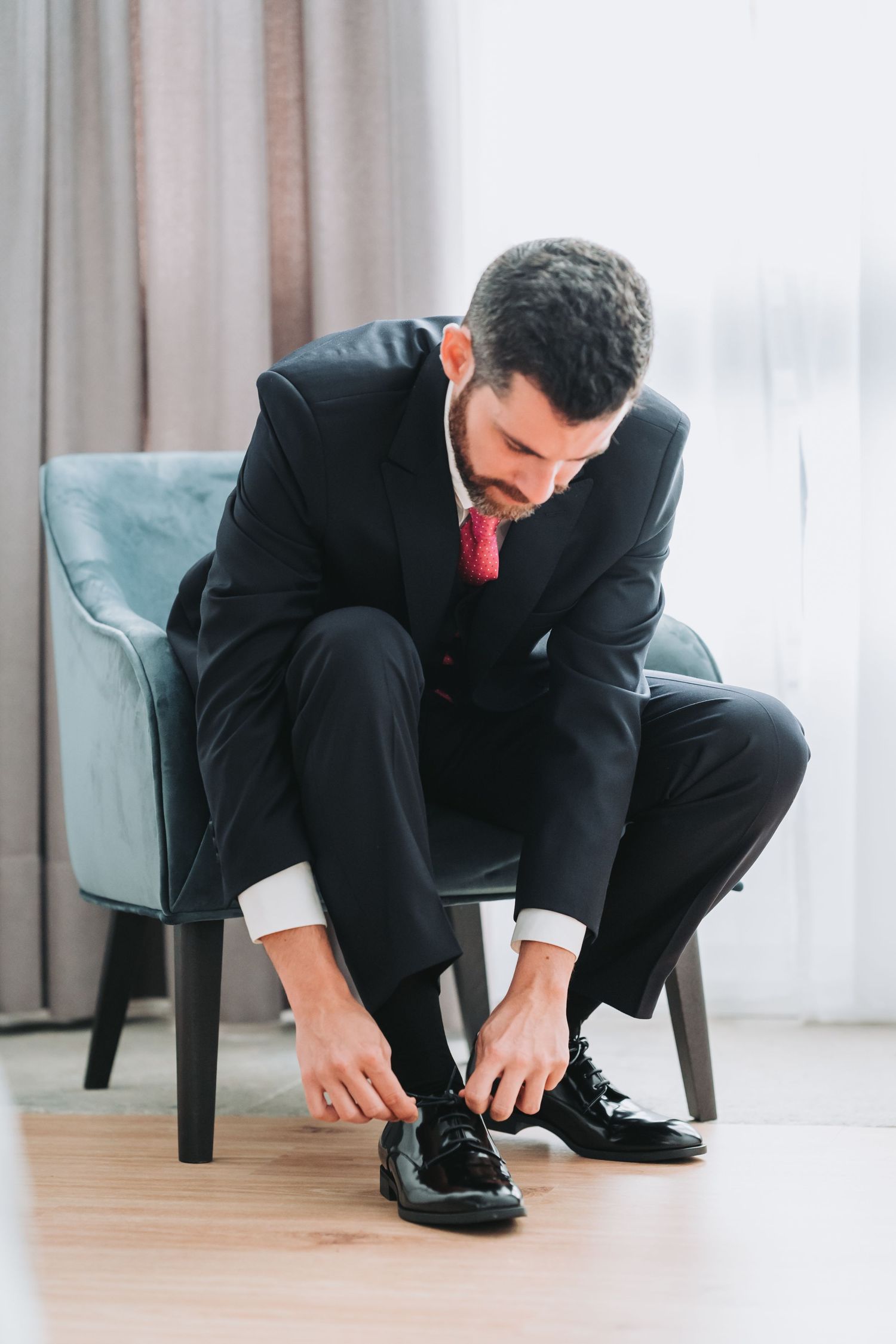 Person in business attire crouching down to tie their polished black dress shoes on a hardwood floor.
