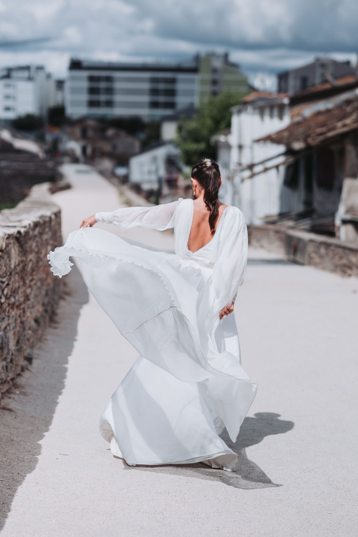 A flowing white wedding dress billows gracefully in the wind against a backdrop of rustic buildings and cloudy skies.