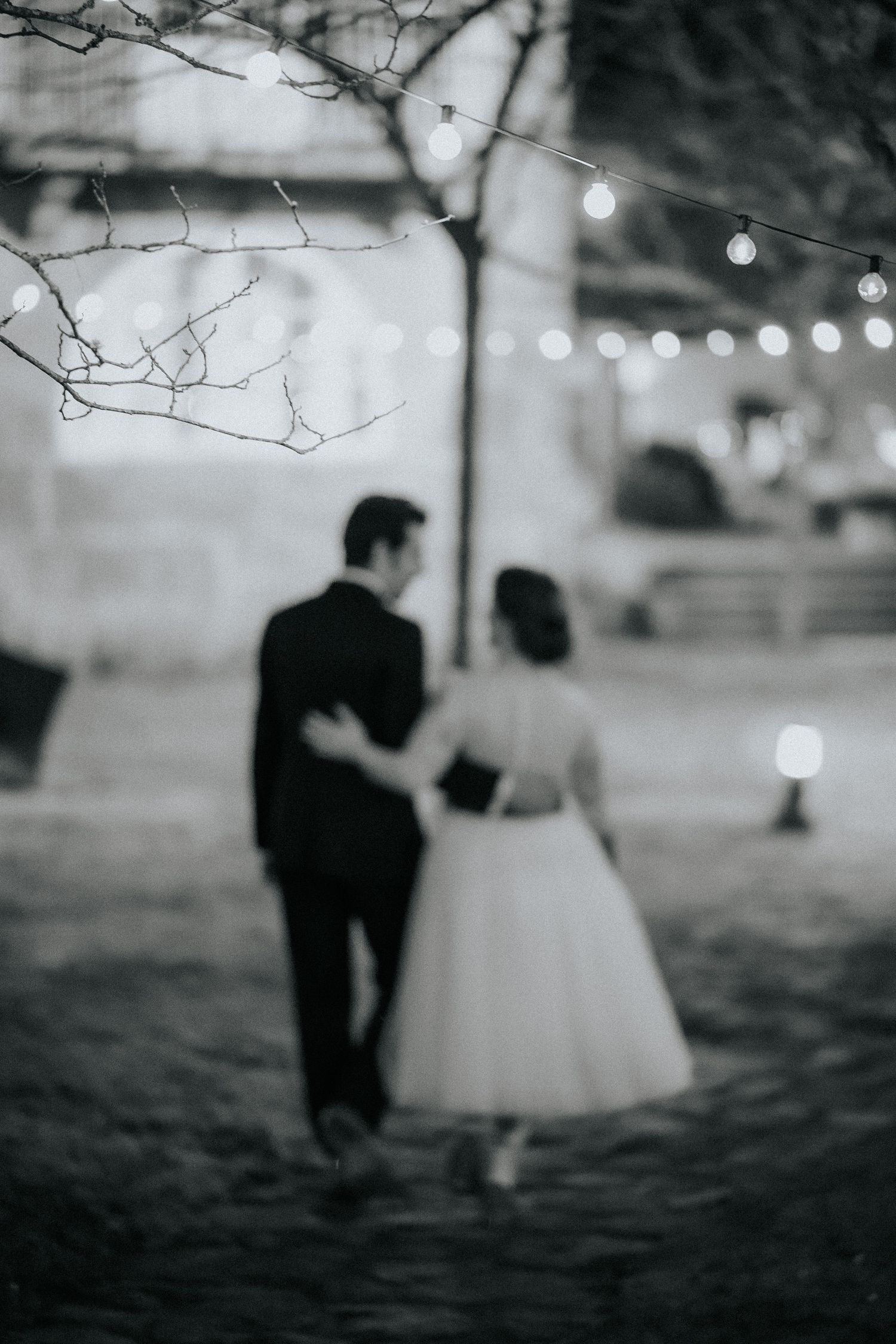 A couple in wedding attire walks through snow at night with twinkling lights in the background.