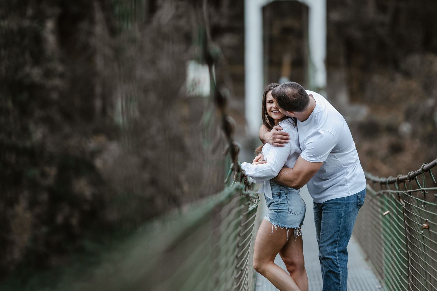 A couple shares a romantic moment on a rustic wooden suspension bridge surrounded by trees.