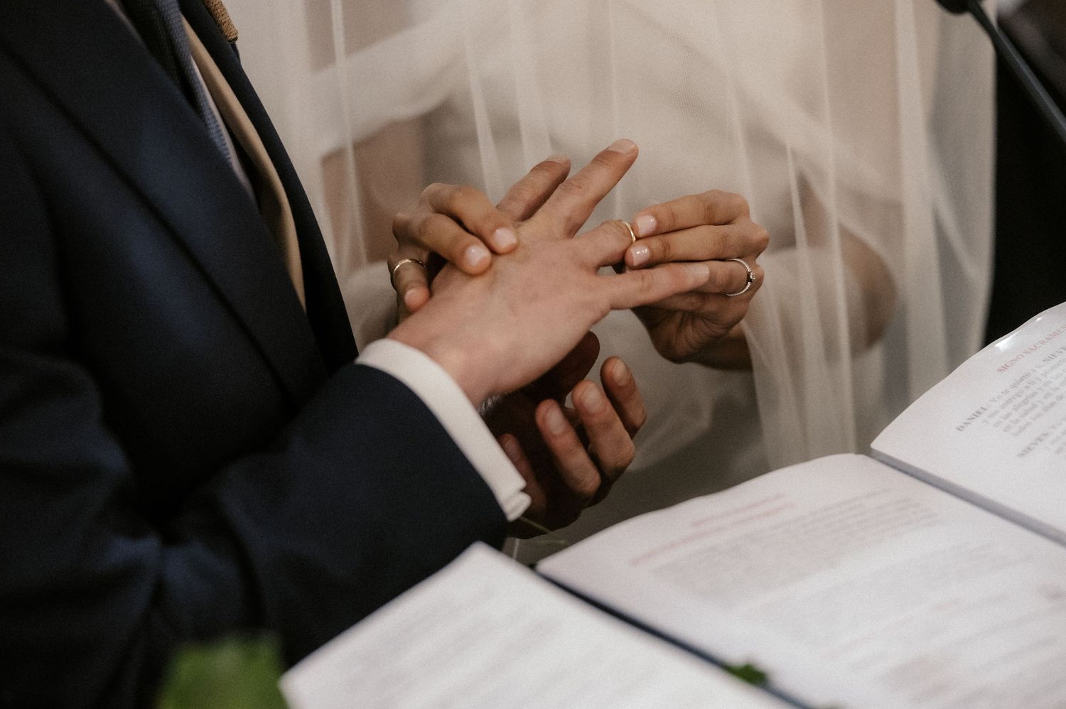 Close-up of hands exchanging wedding rings during ceremony with white veil and open book in frame.
