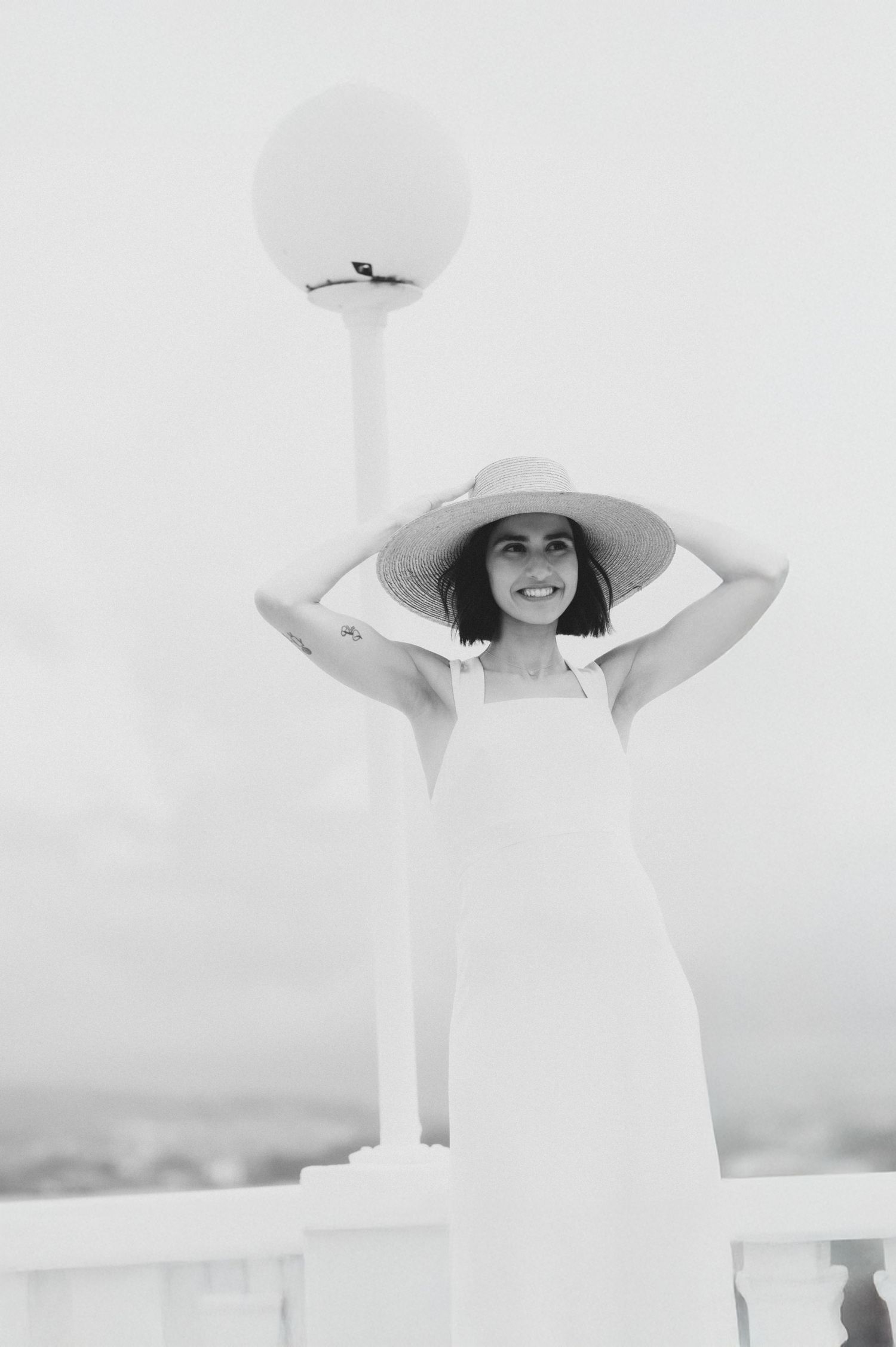 A person in a flowing white dress and wide-brimmed sun hat poses elegantly against a bright background.