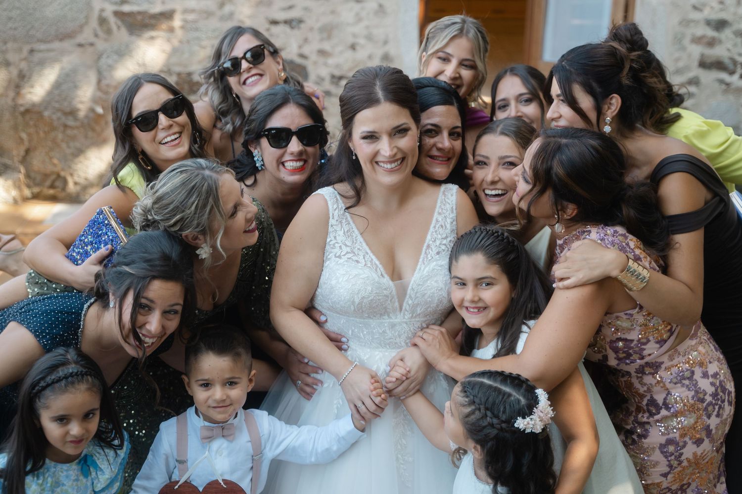 A joyful group photo at a wedding celebration with the bride in a white lace dress surrounded by loved ones outdoors.
