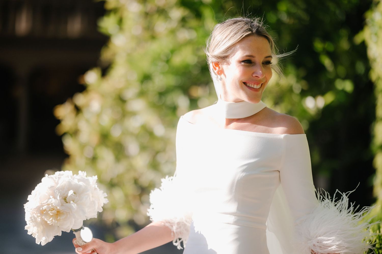 Elegant bride in white gown poses outdoors with flowing veil and floral bouquet in a garden setting.