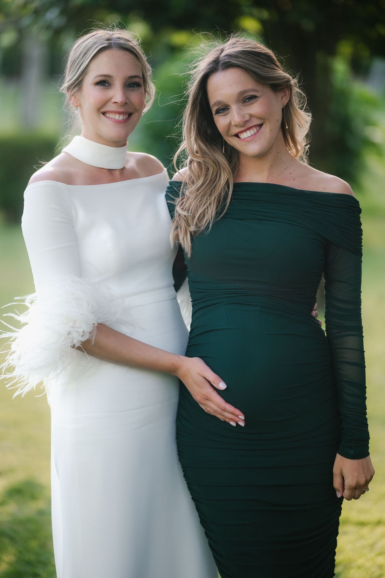 Two women in contrasting white and dark green dresses pose together at an outdoor celebration.