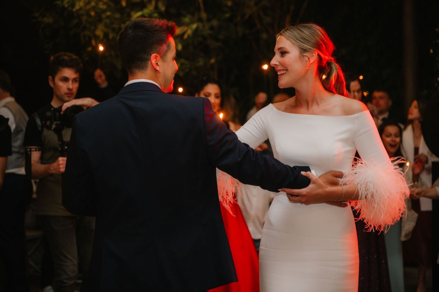 A couple shares their first dance at a wedding reception with warm lighting and guests watching in the background.