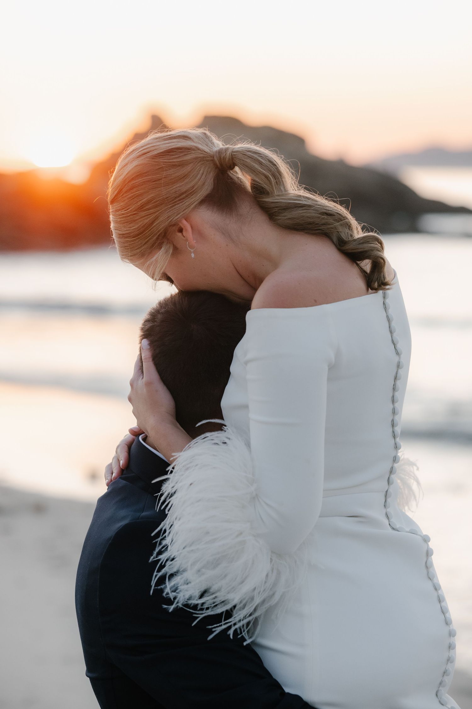 A couple in wedding attire share an intimate embrace at sunset by the water with feather details on the dress.
