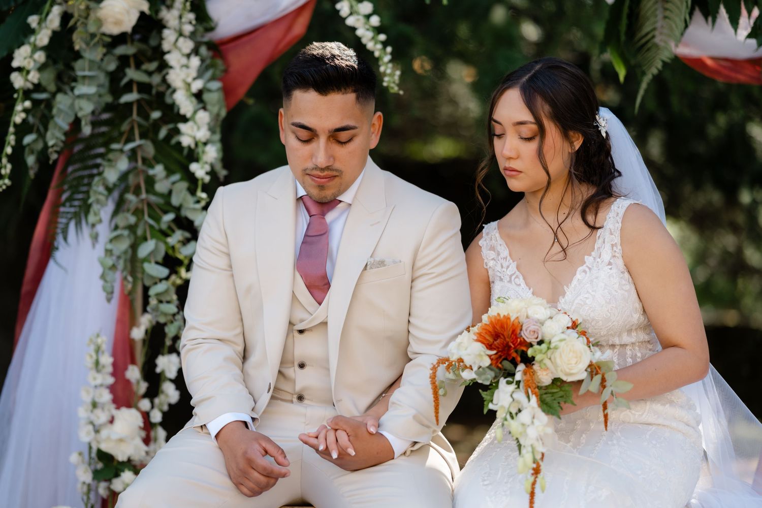 Newlyweds sit together during outdoor garden wedding ceremony with floral arch and red drapery decorations.