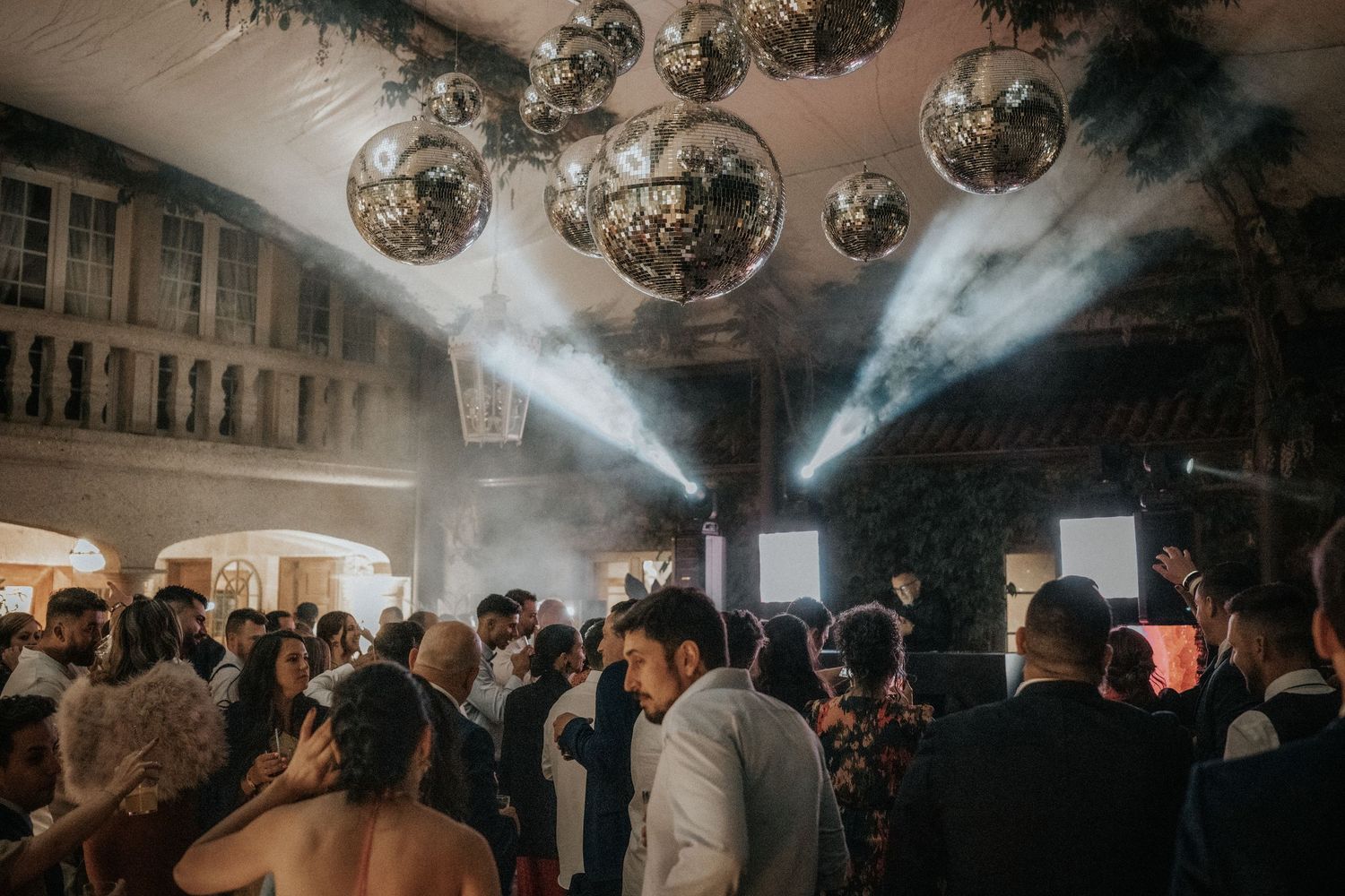 A crowded dance floor at a wedding reception with disco balls overhead and atmospheric smoke effects.