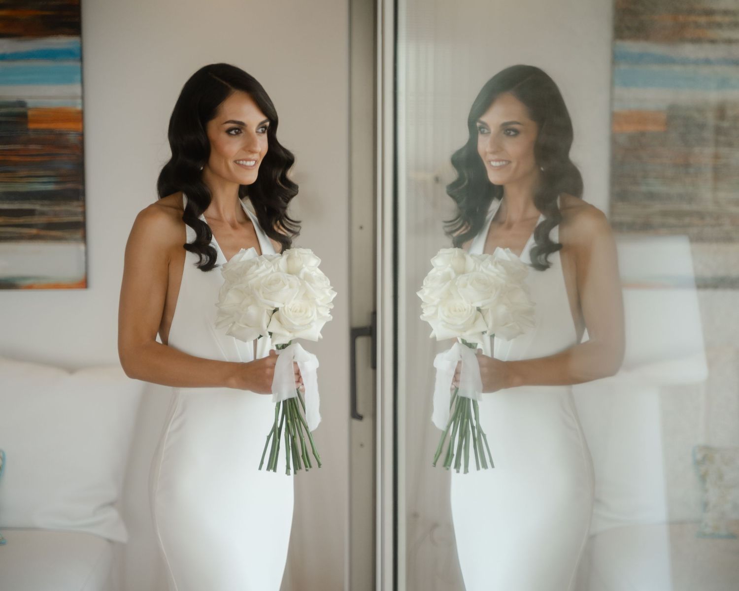 A bride in an elegant white wedding dress holds a bouquet of white flowers while standing in front of a mirror.