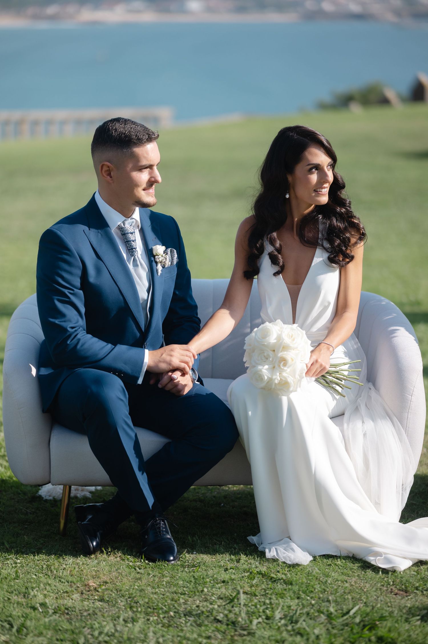Couple sits on white couch during outdoor wedding ceremony with scenic waterfront backdrop.