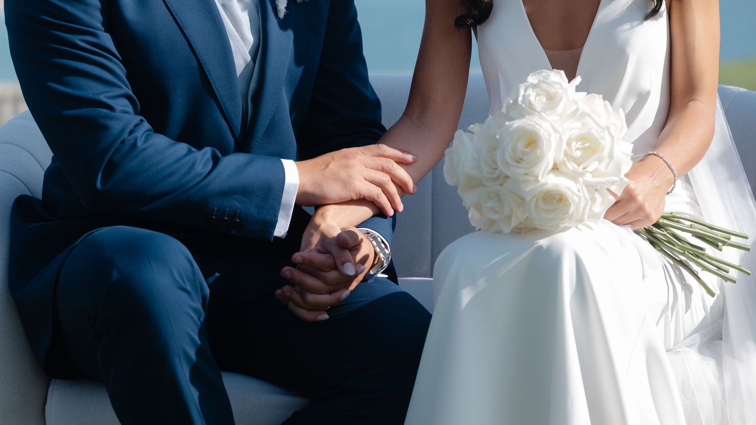 Wedding party members in navy suits and white dresses stand in a line against a white wall for formal photos.