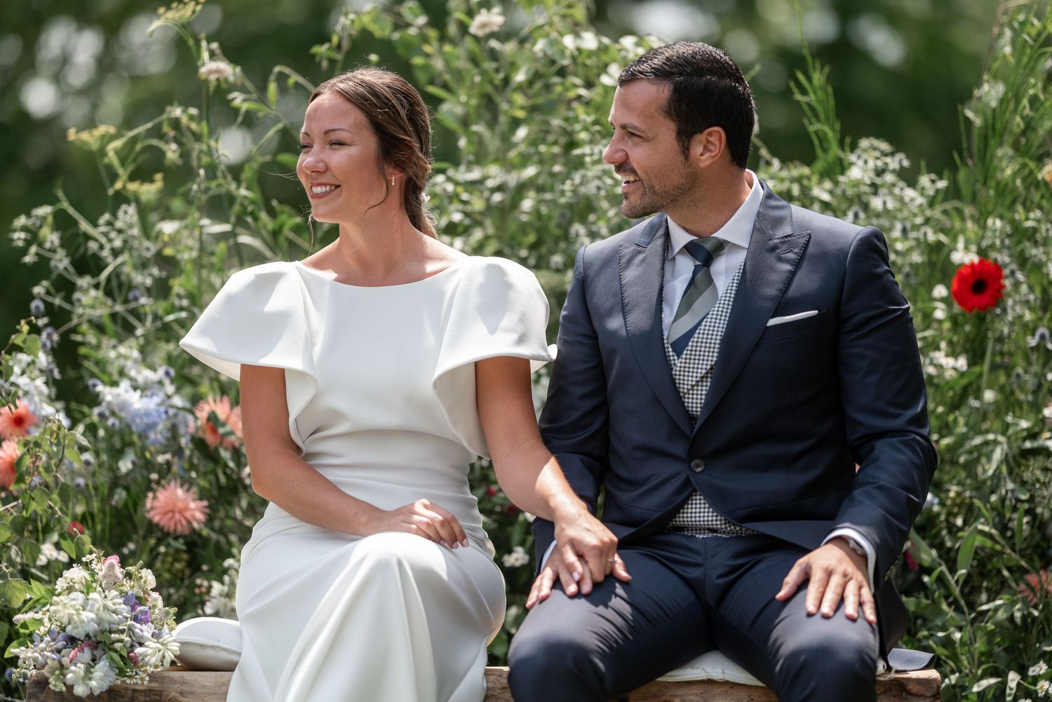 Newlyweds share intimate moments in garden setting surrounded by blooming flowers.