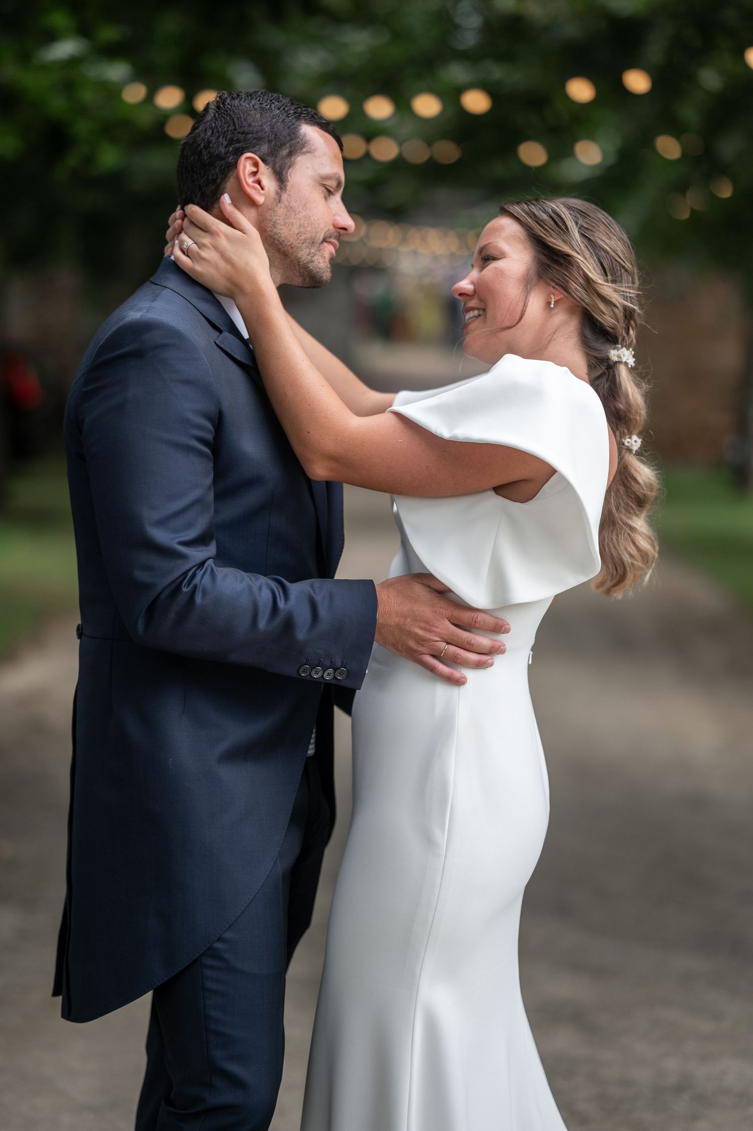 A romantic couple in wedding attire share an intimate moment outdoors with string lights in the background.