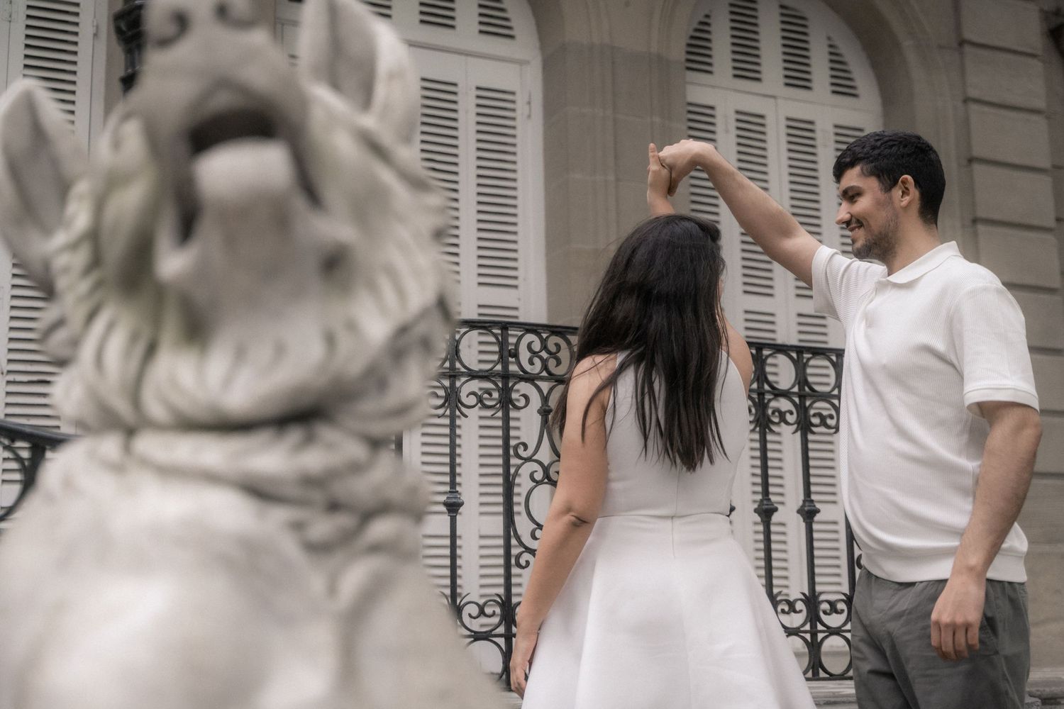 A couple in white outfits poses together on a grand stone balcony with ornate shutters and architectural details.