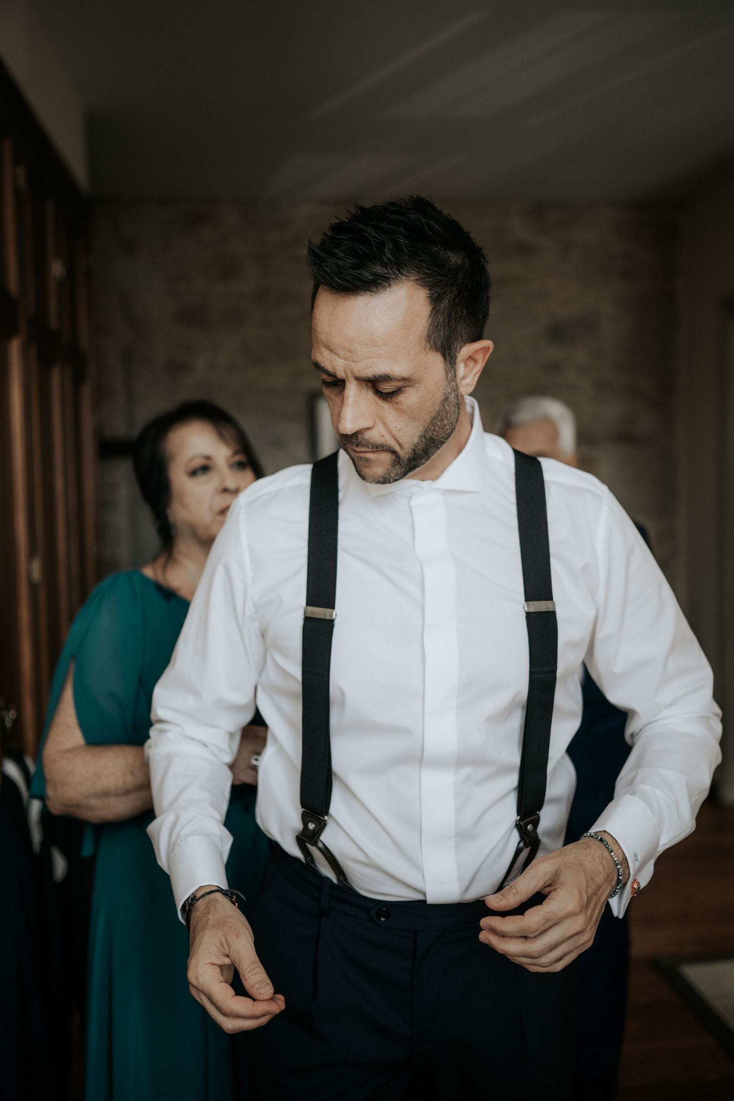 A person in a white dress shirt and black suspenders adjusts their attire while getting ready.
