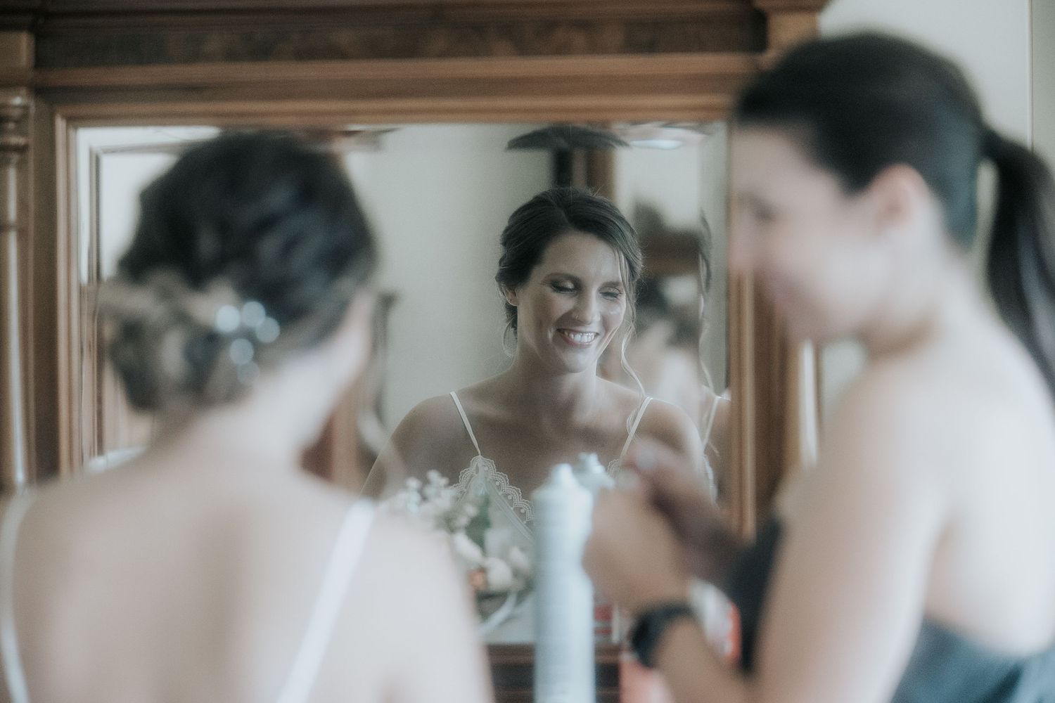 A bride in a white wedding dress gets ready while looking in a mirror during wedding preparations.