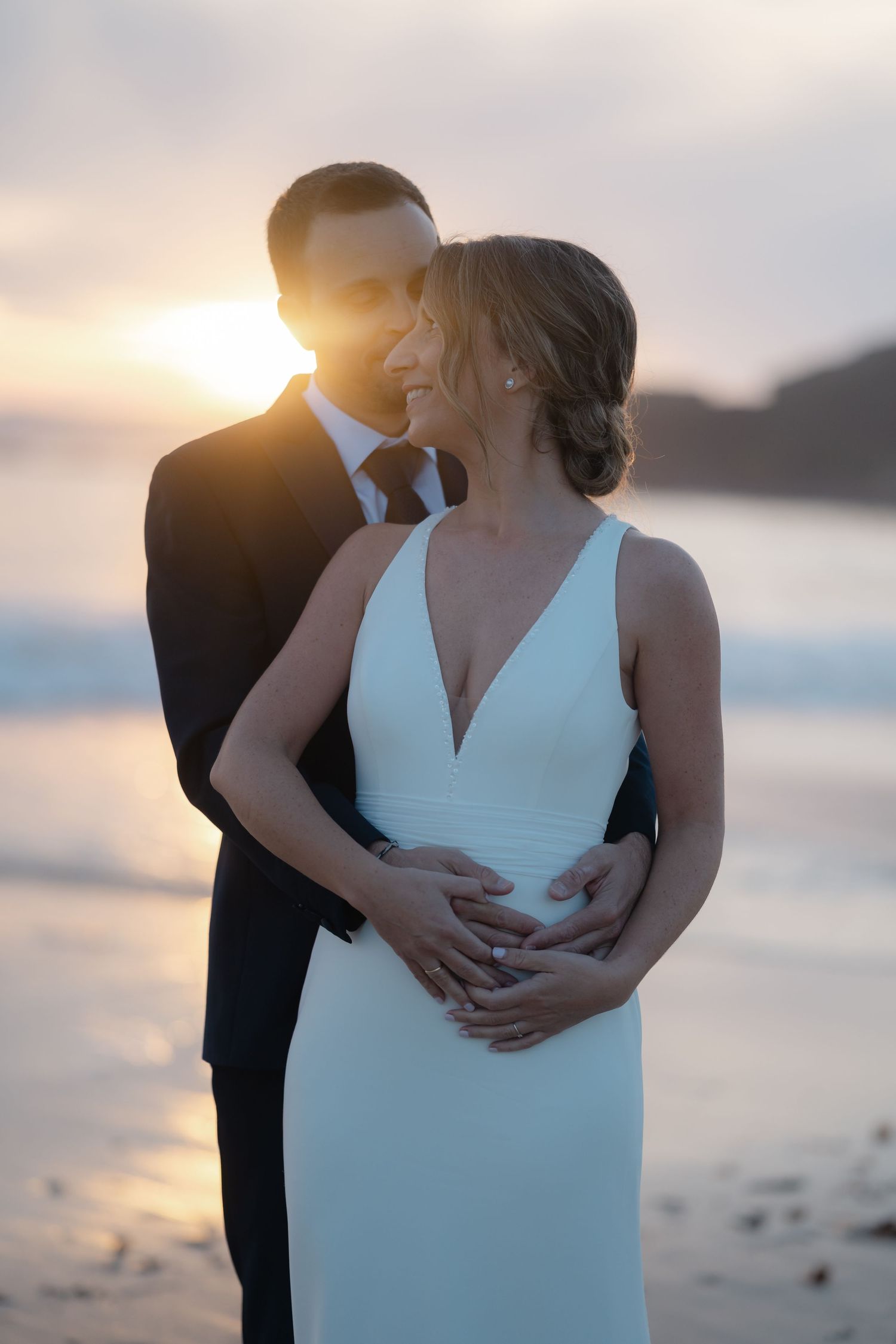 Newlyweds share an intimate moment on a beach at sunset with ocean waves in the background.