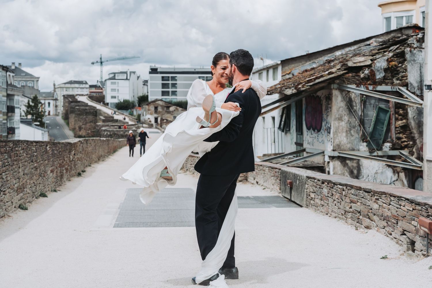 A romantic wedding couple embraces on an old stone wall path with urban buildings in the background.