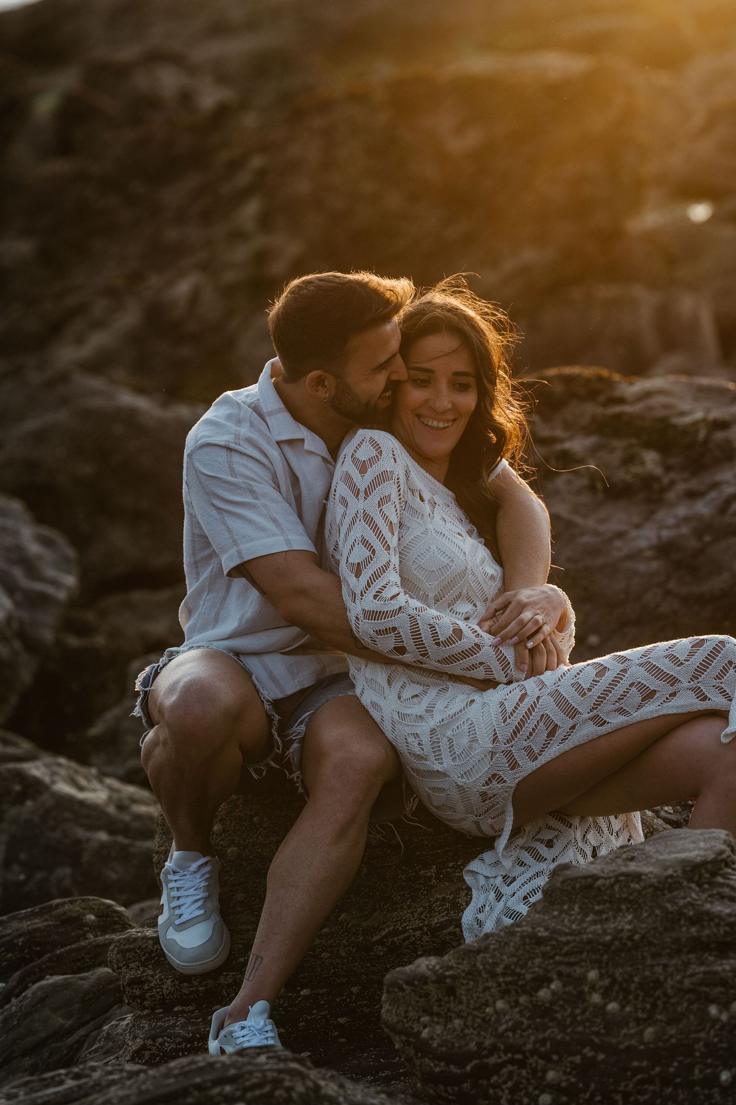 Couple sits intimately on rocky beach during golden hour wearing white lace dress and casual clothing.