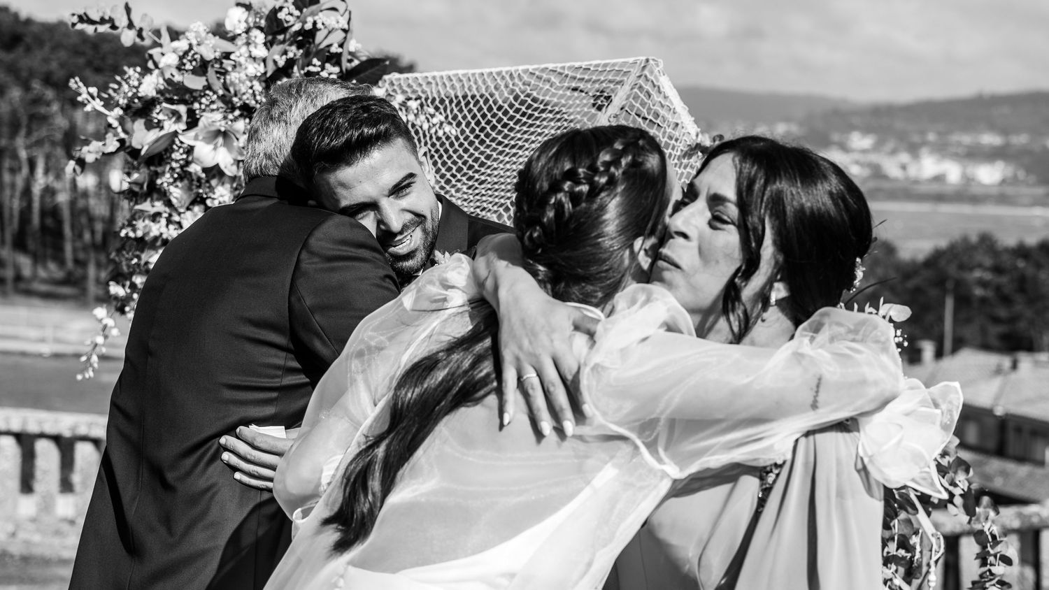 A group of wedding guests share an emotional embrace outdoors with a waterfront view in the background.