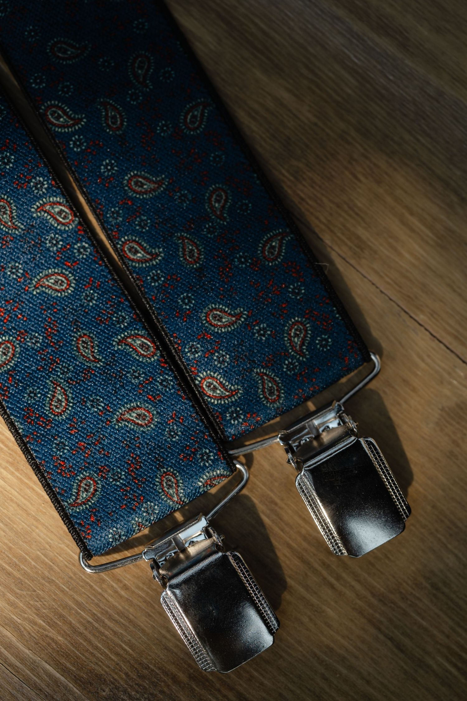 Blue paisley patterned suspenders with silver metal clips on a wooden surface.